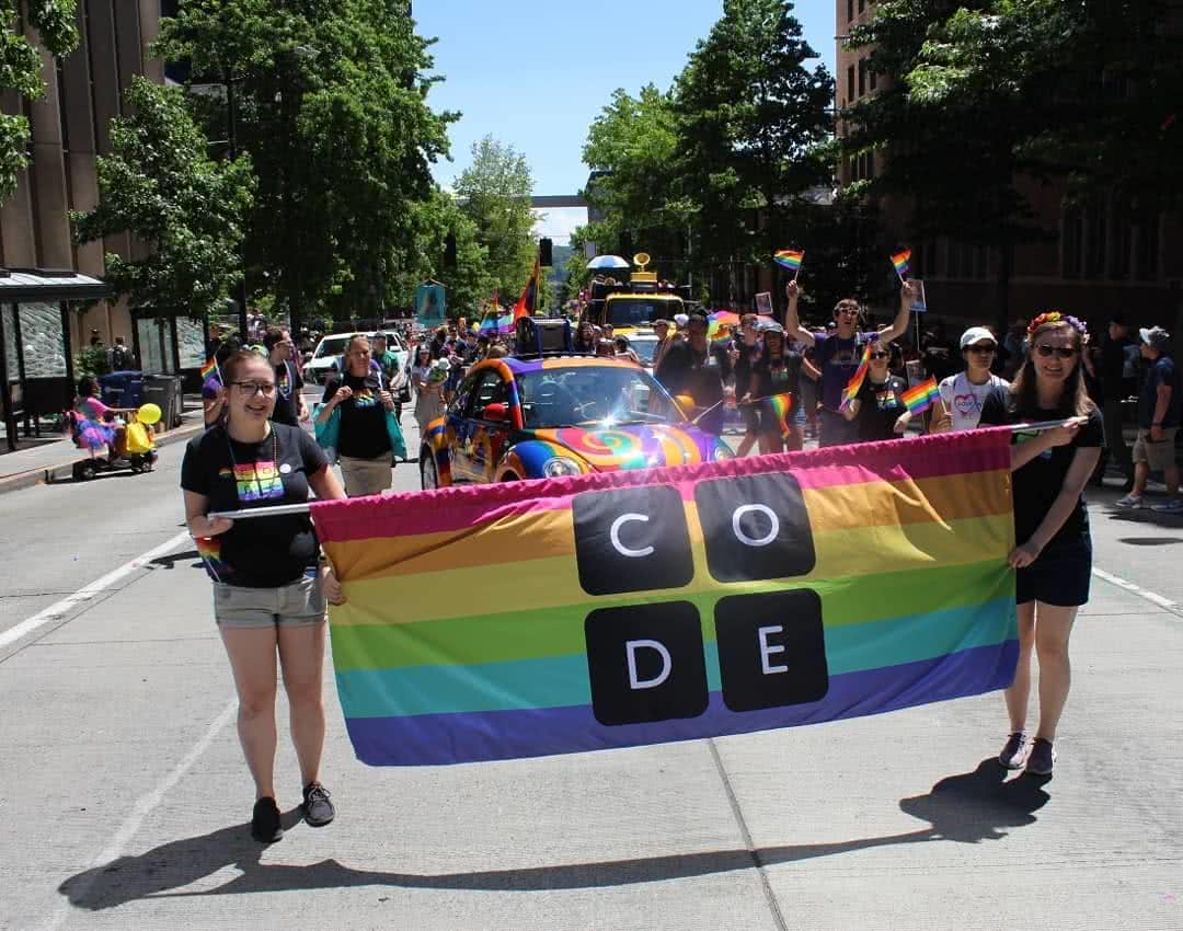 Two feminine-presenting employees smile while holding the ends of a Code.org-branded rainbow banner. Behind them, several people are wearing Code.org shirts and waving rainbow flags while walking alongside a multi-colored Volkswagen bug.